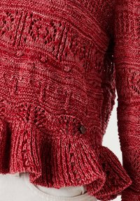Roter Strickpullover mit aufwändigen Mustern und einem gerüschten Saum, getragen über weißen Hosen, mit Fokus auf den Details des Mittelbereichs und der Ärmel.