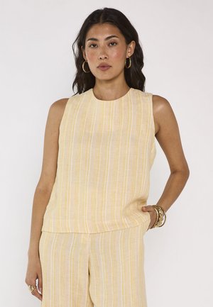 Femme aux cheveux foncés et ondulés, portant un haut sans manches rayé jaune et blanc et un pantalon assorti, accessoirisée avec des boucles d'oreilles créoles dorées et des bracelets.