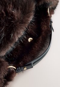 Geantă maro din blană artificială cu o curea netedă din piele neagră. Are o închizătoare cu capse aurie și o textură pufoasă cu lungimi diferite ale firelor de păr.