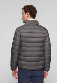 Grå puffjacka med kviltad design och horisontella linjer samt hög krage, visad från baksidan. Slätt material.