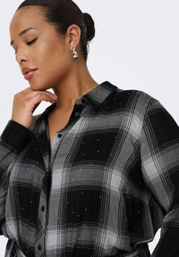 Black and gray plaid shirt with button closure, featuring a collar, subtle embellishments, and a soft fabric texture. Silver hoop earrings.