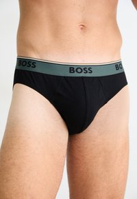 Torse masculin portant un shorty noir avec une ceinture verte portant le mot "BOSS" en lettres noires.