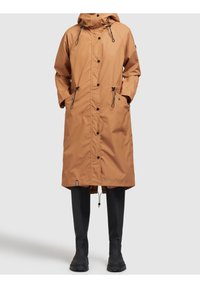 Cappotto impermeabile lungo di colore marrone con cappuccio, cordini regolabili e bottoni a pressione; abbinato a stivali di gomma neri alti fino al ginocchio.