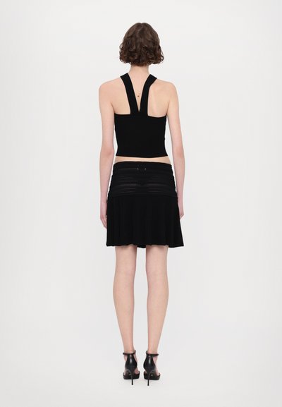 Zwarte crop top met brede banden en open rug, gecombineerd met een gelaagde zwarte geplooide rok. Model draagt zwarte sleehakken.