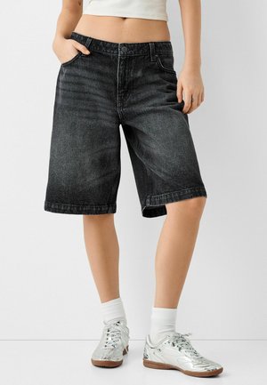 Svarta denimshorts med avslappnad passform, uppvikta fållar och femfickorsdesign, kombinerade med metalliska sneakers och vita ankelsockor.
