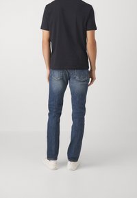 Mörkt T-shirt, blå denimjeans med rak passform, blandad bomullsmaterial och vita sneakers, sedd bakifrån mot en enkel bakgrund.