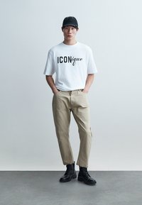 Νέος άνδρας που φοράει λευκό μπλουζάκι με την επιγραφή "ICONique", μπεζ παντελόνι, μαύρα στρατιωτικά μποτάκια και μαύρη καπέλο μπέιζμπολ, στέκεται με τα χέρια στις τσέπες.