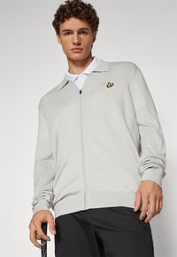 Light gray zip-up cardigan with a collar, made from textured fabric. Features a small yellow logo and long sleeves. Worn over a white polo.