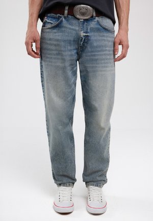 Jean en denim bleu clair avec une coupe décontractée, une finition délavée, cinq poches et une boucle de ceinture décorative en argent. Portés avec des baskets blanches.