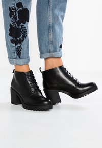 Botas de cuero negro hasta el tobillo con parte superior texturizada, punteras redondas, cordones y tacones altos y gruesos. Combinadas con jeans de color azul claro que presentan bordados florales.