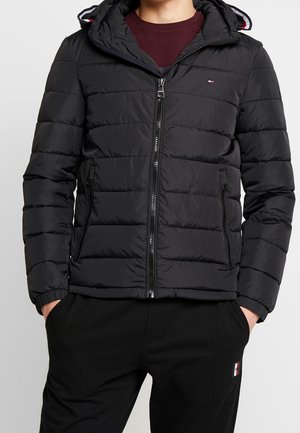 Person iført en sort quiltet hættetrøje over en burgunderfarvet skjorte, med hænderne i lommerne på sorte bukser, vist fra nakke til hofter.