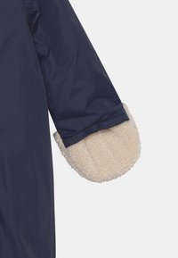 Manica di un impermeabile blu navy con un morbido polsino in pile color crema. Il polsino presenta un aspetto texturizzato e soffice, in contrasto con l'esterno liscio.