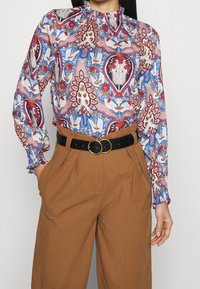 Blusa a maniche lunghe con un motivo paisley multicolore in blu, rosso e rosa. Presenta un collo alto e polsini arricciati. Abbinata a pantaloni marroni.