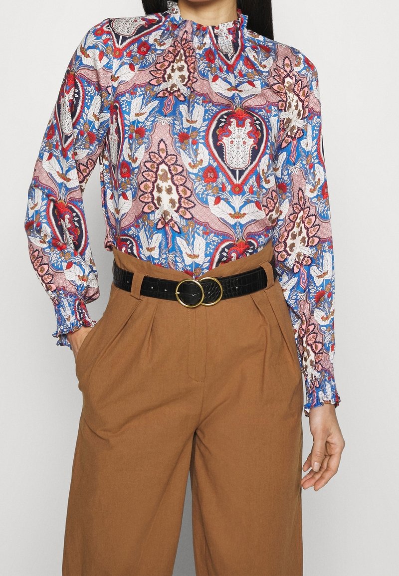 Blusa a maniche lunghe con un motivo paisley multicolore in blu, rosso e rosa. Presenta un collo alto e polsini arricciati. Abbinata a pantaloni marroni.