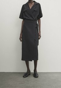 Modell trägt ein schwarzes Wickelkleid mit kurzen Ärmeln, tailliertem Design und schwarzen geschlossenen Schuhen, steht vor einer schlichten Wand.