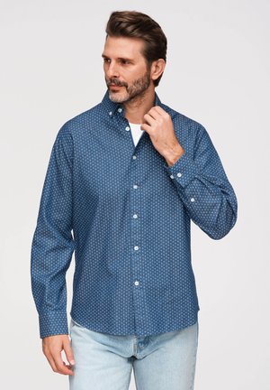 Mężczyzna z krótkimi brązowymi włosami i brodą, ubrany w niebieską wzorzystą koszulę zapinaną na guziki oraz jasnoniebieskie dżinsy, poprawia kołnierz prawą ręką.