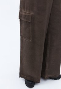 Bruna vida-byxor i cargo-stil med en texturerad tyg, som har en stor sidficka och synliga sömmar. Bärs med svarta kraftiga stövlar.