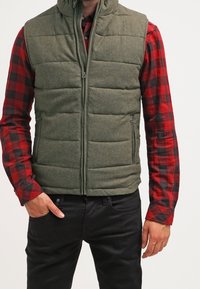 Olivgrüne gepolsterte Weste mit hohem Kragen, Frontreißverschluss und zwei Seitentaschen. Getragen über einem rot-schwarz karierten Flanellhemd.