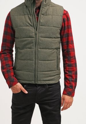 Zielono-oliwkowa kamizelka ocieplana z wysokim kołnierzem, zamkiem błyskawicznym z przodu i dwiema bocznymi kieszeniami. Noszona na czerwoną i czarną koszulę w kratę flanelową.