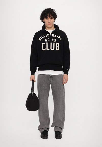 Μαύρη μπλούζα φούτερ με την επιγραφή "Billionaire Boys Club" σε κρέμα απόχρωση, συνδυασμένη με γκρι παντελόνι τύπου wide-leg και μαύρα παπούτσια, κρατώντας μια μικρή μαύρη τσάντα.