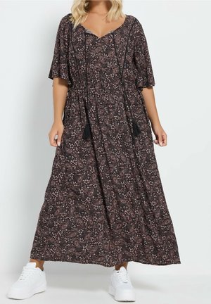 Kvinde iført en brun paisley maxi-kjole med flagrende ærmer og hvide sneakers, stående mod en ensfarvet lys baggrund.