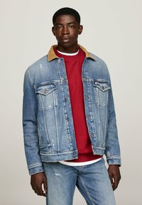 Chaqueta de mezclilla con un cuello de pana color beige, de color azul claro, con bolsillos frontales y detalles desgastados, usada sobre un sudadero rojo.