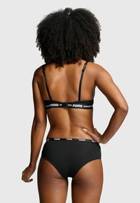 Frau mit lockigem Haar, die ein schwarzes Puma-Bra und Unterwäsche-Set trägt, von hinten vor einem schlichten Hintergrund gezeigt.