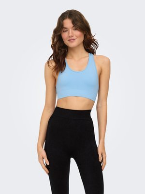 Woman with wavy brown hair wearing a light blue sports bra and high-waisted black leggings, standing against a plain light background.