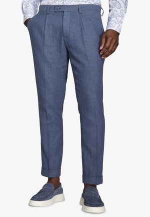 Mann trägt blaue, strukturierte Anzughose mit Umschlagsäumen, kombiniert mit blauen Veloursleder-Slippern und einem weißen Hemd mit Blumenmuster.