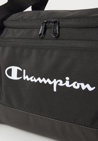 Svart nylonveske med dobbel glidelås, med en fremtredende hvit "Champion"-logo, teksturert overflate og strukturert design.