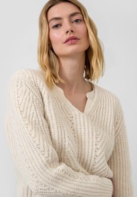Beige Strickpullover mit tiefem V-Ausschnitt, strukturiert mit gerippten Mustern und langen Ärmeln. Weiches Material mit dezenten Glitzereffekten.