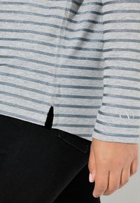 Graues Langarmoberteil mit horizontalen blauen Glitzerstreifen, das am Saum einen seitlichen Schlitz hat. Getragen mit schwarzen Jeans.
