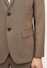 Καφέ καρό σακάκι με δύο μαύρα κουμπιά, τσέπη με καπάκι και ταιριαστό παντελόνι, φορεμένο από ένα άτομο με ορατό χέρι στο πλάι.