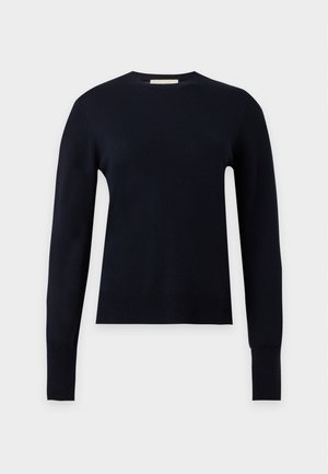 Donna che indossa una maglia nera a maniche lunghe, gonna grigia alla caviglia e ballerine nere, in piedi su uno sfondo chiaro e uniforme.
