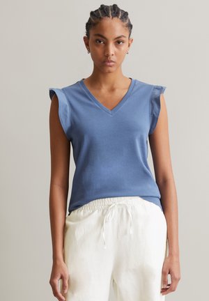 Blaues V-Ausschnitt ärmelloses Top mit weichen Rüschensäumen, aus leichtem Stoff; kombiniert mit weißen Hose mit elastischem Bund, glatte Textur.