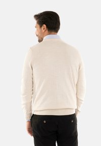 Maglione lavorato a maglia beige con maniche lunghe e scollo rotondo, abbinato a pantaloni scuri con dettagli in bottone sulle tasche posteriori.