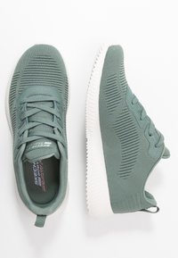 Grüne Strick-Sneaker mit strukturiertem Obermaterial, weißer Sohle, Schnürdesign und einem Etikett, das "Skechers Memory Foam" innen zeigt.