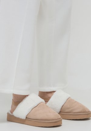 Babouches en daim ouvertes à l'arrière de couleur beige, avec une bride en fausse fourrure blanche et une semelle texturée. Les babouches ont une finition douce et un bout arrondi.