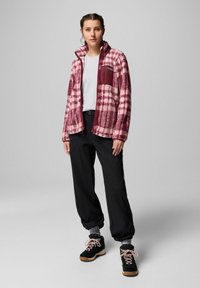 Giovane donna che indossa una giacca di pile a quadretti rosa, una maglietta bianca, pantaloni neri e scarpe da trekking nere con lacci rosa, in piedi contro uno sfondo neutro.