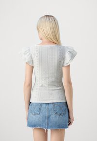 White cotton top with short ruffled sleeves, featuring eyelet patterns and a form-fitting design. Worn with a denim mini skirt.