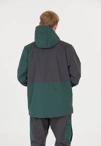Veste imperméable avec capuche, présentant une partie supérieure gris foncé et une partie inférieure verte. Poches zippées et coupe décontractée.