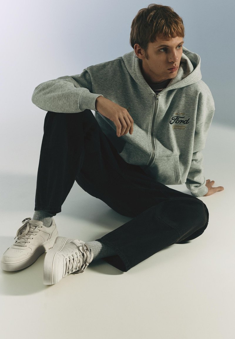 Jeune personne assise par terre, portant un sweat à capuche gris Ford, un pantalon noir, des chaussettes grises et des baskets blanches, regardant sur le côté.