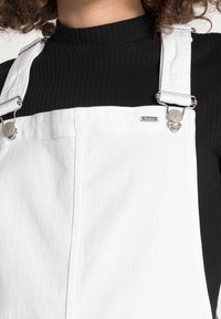 Vita denimoveraller med silverfärgade detaljer, justerbara axelband och en liten varumärkesetikett. Bärs över en svart ribbad långärmad tröja.