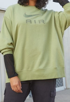 Person trägt übergroßen hellgrünen Nike Air Sweatshirt und schwarzes, geschichtetes Mesh-Ärmeltop mit schwarzen Cargo-Hosen im Freien.