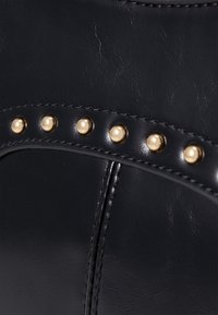 Sac à main en cuir synthétique noir avec une finition brillante, des clous décoratifs dorés et des coutures élégantes le long des coutures.