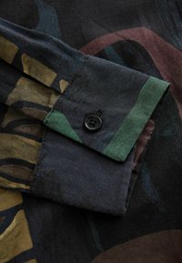 Dettaglio di una camicia di cotone su uno sfondo scuro con motivi astratti in verde, marrone e bordeaux. Presenta un bottone nero sul polsino.