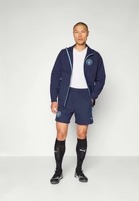 Sötétkék sport melegítő világoskék díszítéssel, cipzáras kabáttal, rövid ujjú pólóval és hozzáillő rövidnadrággal. Fekete focizokni és stoplis cipő.