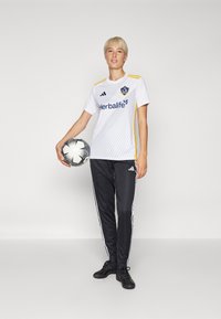 Balta Adidas krekls ar dzeltenām akcentiem un diagonālām svītrām, melnas Adidas sporta bikses ar baltām sānu svītrām, turēdams melnbaltu futbola bumbu.