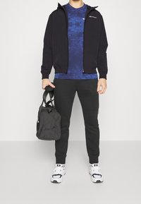 Black zip-up hoodie, blue patterned T-shirt, black joggers, white sneakers with black accents, and a black duffel bag.
