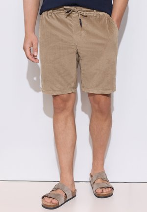 Mand iført beige kordfløjls shorts med snøre i taljen og taupe sandaler med dobbelt strop, stående mod en ensfarvet hvid baggrund.